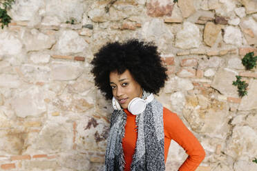 Young woman with headphones and scarf standing against stone wall on sunny day - XLGF01065