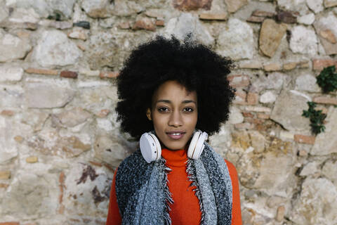 Junge Frau mit Kopfhörern in warmer Kleidung an einer Steinmauer, lizenzfreies Stockfoto