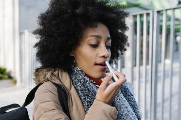 Afro young woman applying lipstick while standing on street - XLGF01068