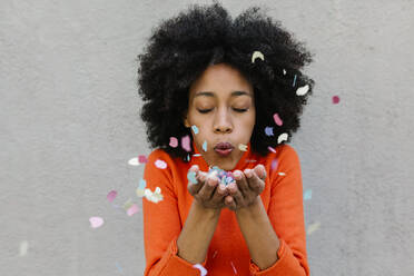 Afro young woman with eyes closed blowing confetti against wall - XLGF01072