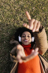 Lächelnde Frau hört Musik und gestikuliert, während sie im Park im Gras liegt - XLGF01076