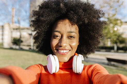 Lächelnde Afro-Frau mit Kopfhörern macht ein Selfie im Park an einem sonnigen Tag - XLGF01078