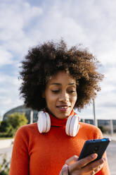 Afro-Frau mit Kopfhörern, die ein Mobiltelefon gegen den Himmel benutzt - XLGF01081