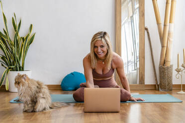 Smiling blond mature woman using laptop while doing yoga in living room - DLTSF01607