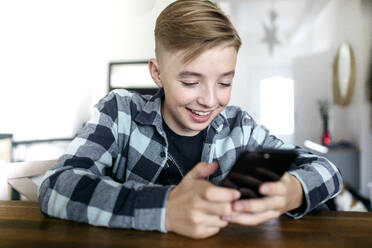 Smiling boy using smart phone while sitting at table in living room - KMKF01483