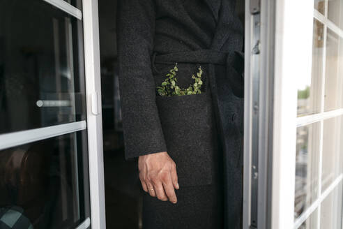 Älterer Mann in schwarzem Mantel mit Pflanze in der Tasche steht vor der Tür - KMKF01485