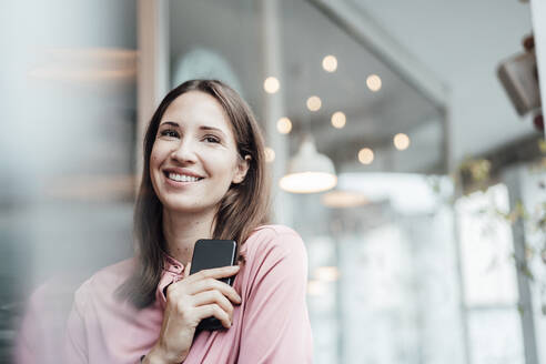 Lächelnde Freiberuflerin, die ihr Smartphone in der Hand hält und im Café wegschaut - JOSEF03102