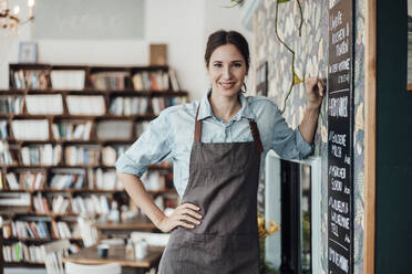 Smiling female owner with hand on hip leaning by wall at coffee shop - JOSEF03119