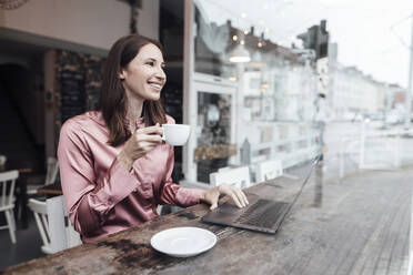 Happy businesswoman with coffee cup working on laptop at table in cafe - JOSEF03150