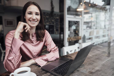 Lächelnde Unternehmerin mit Kaffeetasse und Laptop am Tisch in einem Cafe - JOSEF03151
