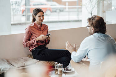 Geschäftsfrau hält ihr Smartphone in der Hand, während sie mit einem männlichen Kollegen im Café diskutiert - JOSEF03190