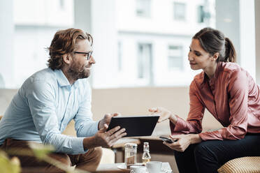 Smiling male and female colleagues discussing over digital tablet during meeting in coffee shop - JOSEF03191