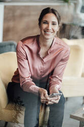 Happy businesswoman with smart phone sitting on sofa in coffee shop - JOSEF03222