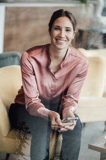 Glückliche Geschäftsfrau mit Smartphone auf dem Sofa in einem Café sitzend - JOSEF03222
