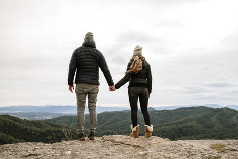 Couple holding hands while standing at viewpoint looking at mountains against sky - AFVF08141