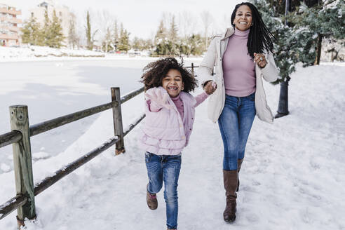 Happy mother and daughter holding hands while running in park during winter - EBBF02303