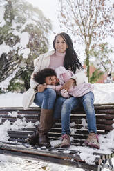 Daughter resting head on mother's lap while sitting on bench in park during winter - EBBF02308