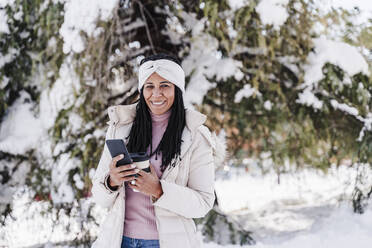 Lächelnde Frau mit Smartphone und Kaffeetasse im Park im Winter - EBBF02310