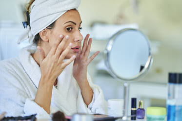 Frau beim Eincremen ihres Gesichts vor einem Spiegel zu Hause - KIJF03531