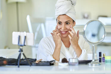 Smiling female influencer applying face cream on her face while blogging through mobile phone at home - KIJF03533