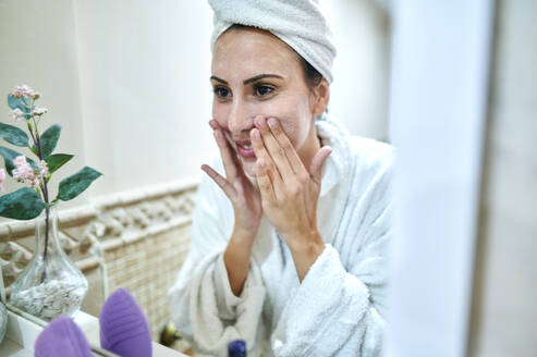 Spiegelung einer lächelnden Frau, die im Badezimmer Gesichtscreme aufträgt - KIJF03544