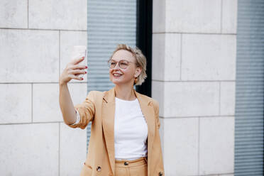 Smiling mature female professional taking selfie through smart phone against wall - KMKF01488