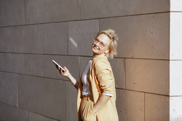 Smiling female entrepreneur with smart phone standing against wall - KMKF01489