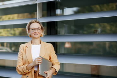 Selbstbewusste reife Geschäftsfrau mit Laptop vor einem Bürogebäude - KMKF01502