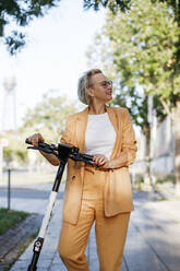 Smiling mature female commuter looking away while standing with electric push scooter on footpath - KMKF01505