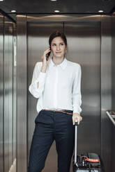 Businesswoman with suitcase taking on smart phone while standing in elevator - JOSEF03261