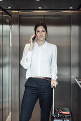 Geschäftsfrau mit Koffer, die im Aufzug stehend ihr Smartphone benutzt - JOSEF03261