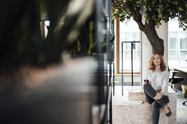 Female entrepreneur with smart phone sitting on retaining wall in office - JOSEF03268