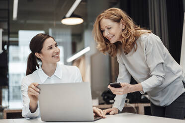 Smiling female colleagues discussing over laptop at desk in office - JOSEF03269