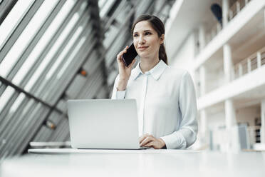 Lächelnde Unternehmerin, die mit einem Smartphone telefoniert, während sie im Korridor an einem Laptop arbeitet - JOSEF03276