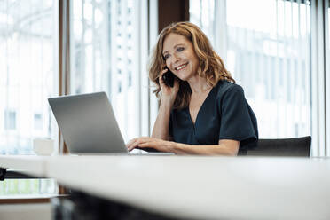 Smiling female entrepreneur working on laptop while talking over smart phone at board room - JOSEF03278