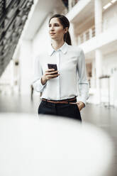 Confident businesswoman with smart phone looking away while standing in corridor - JOSEF03280