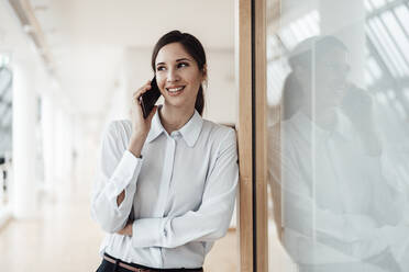 Lächelnde Geschäftsfrau, die mit ihrem Smartphone spricht, während sie sich im Büro an die Wand lehnt - JOSEF03292