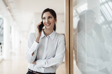 Smiling businesswoman talking on smart phone while leaning by wall in office - JOSEF03292