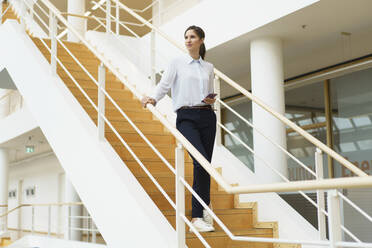 Female entrepreneur with smart phone standing on steps in office corridor - JOSEF03301