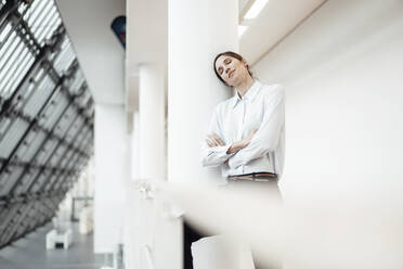 Female entrepreneur with eyes closed leaning by wall in corridor at office - JOSEF03314