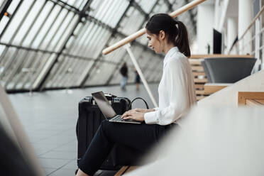 Businesswoman working on laptop while sitting in office corridor - JOSEF03318