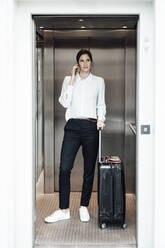 Female entrepreneur with suitcase taking on smart phone while standing in elevator - JOSEF03331