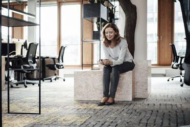 Unternehmerin, die ihr Smartphone benutzt, während sie auf einer Stützmauer im Büro sitzt - JOSEF03345