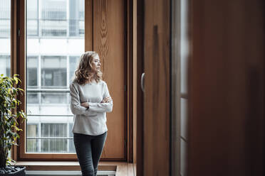 Female entrepreneur with arms crossed looking through window in office - JOSEF03346