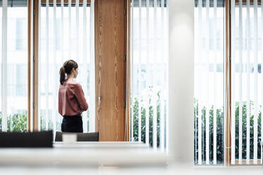 Businesswoman looking through window while standing in office - JOSEF03413