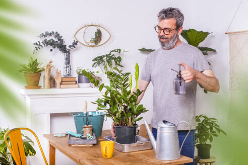 Man spraying water on Zamioculcas Zamiifolia potted plant while standing at home - RTBF01517