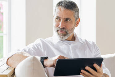 Geschäftsmann mit digitalem Tablet, der wegschaut, während er zu Hause sitzt - SBOF02544