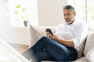 Businessman using mobile phone while sitting on sofa at home - SBOF02549