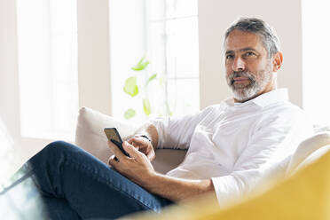 Businessman with smart phone looking away while sitting at home - SBOF02550