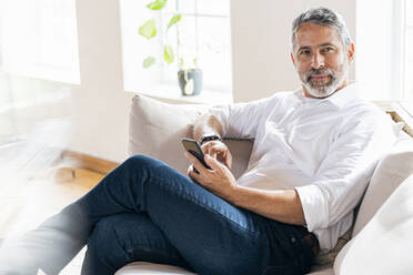 Businessman with mobile phone looking away while sitting at home - SBOF02551